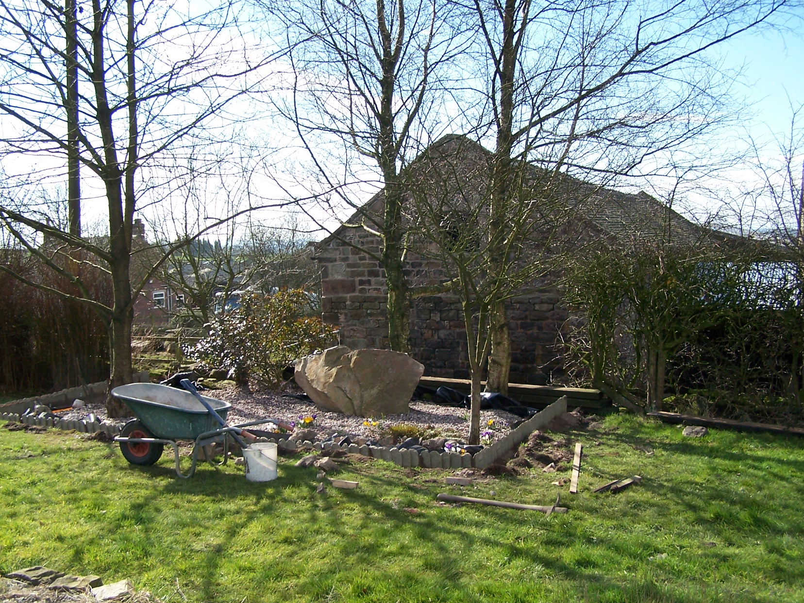 wheelbarrow on lawn in front of gravel bed with boulder and trees